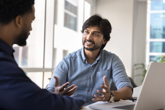 Indian businessman leads formal conversation with company client met in office seated at desk with laptop, sharing current project details, engaged in collaboration planning. Business communication