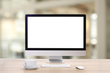 Computer monitor with blank screen and cup on wooden desk against blurred background. Mockup for design
