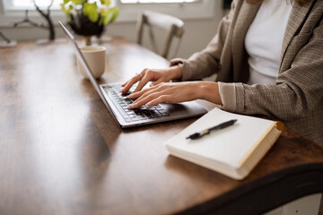 Person writing in notebook beside laptop on wooden desk.