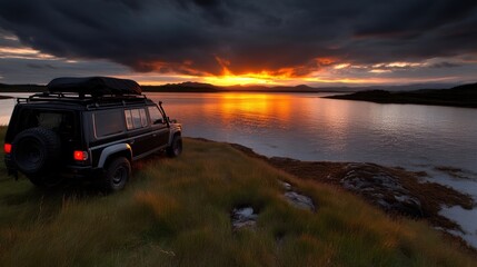 Fototapeta premium Rugged SUV at sunset by a tranquil lake. A powerful vehicle rests on a grassy knoll overlooking a serene lake, bathed in the warm glow of a dramatic sunset