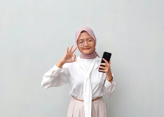 Fototapeta premium Smiling Young Muslim Woman in Hijab Showing OK Hand Gesture While Holding Smartphone Against Plain Background