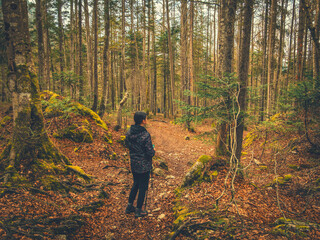 A teenage girl walks alone through a vibrant forest near Kranjska Gora, Slovenia. The path is surrounded by lush evergreen trees and the sky is overcast, creating a moody yet colorful atmosphere.