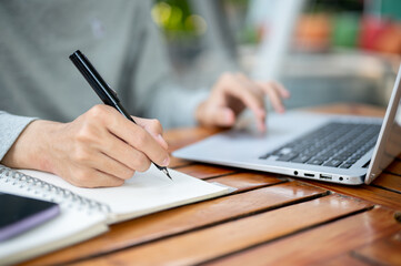 A hand holding a pen writing in notebook and other hand tapping on laptop sitting at wooden table.