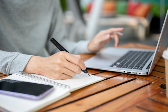 A hand holding a pen writing in notebook and other hand hovering over laptop sitting at wooden table