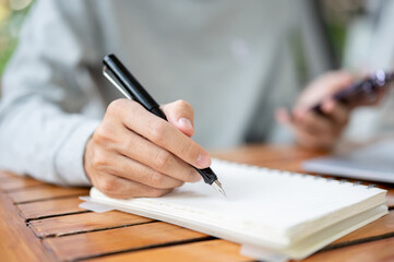 Close up of a hand holding pen writing in notebook and other hand holding smartphone. Study outdoor.
