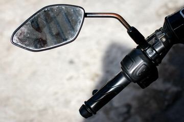 Close up of a motorcycle's left handlebar and side mirror, showing the details of the throttle grip, control switches, and a slightly rusted mirror stem, under natural daylight