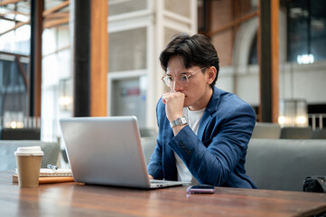 Fototapeta premium Blue suit asian man is looking intently at his laptop on table while sitting in cafe or coffee shop.