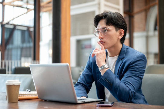 Blue suit asian man thinking thoughtfully in front of a laptop while sitting in cafe or coffee shop.