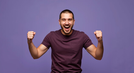 Naklejka premium Joyful young man with short hair in purple t-shirt smiling and raising fists on purple background. Excitement, victory, happiness. Clothing ads, motivation, celebration, psychology, copy space