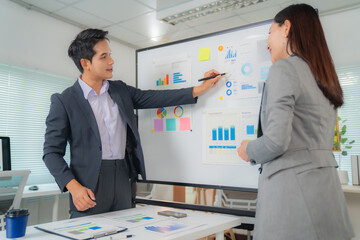 Two asian businesspeople in suits are discussing company performance and analyzing charts and graphs on a whiteboard during a meeting in a modern office