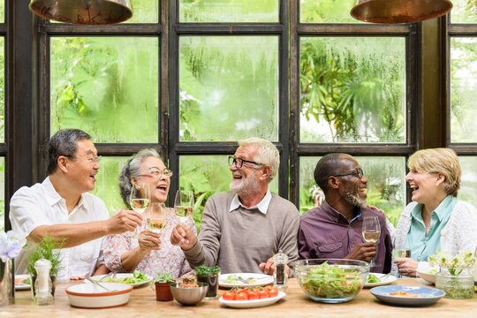 Group of diverse seniors enjoying a meal together, laughing and toasting with wine. Multicultural gathering of elderly friends sharing a joyful moment. Happy diverse senior people at dinner. - Powered by Adobe