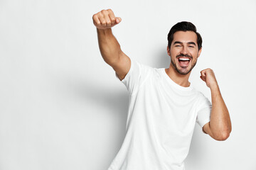 Happy cheerful young man wearing a white t-shirt, smiling widely, raising his fist in excitement, isolated on a plain light background. Joyful and energetic concept.