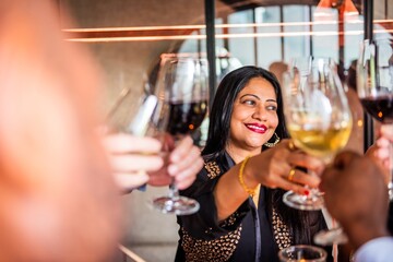 Woman celebrating with friends, holding wine glass, smiling. Festive gathering, diverse group, joyful atmosphere. Cheers with wine, happy celebration. Indian woman at dinner party.