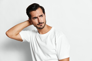Thoughtful young man in a casual white t-shirt looking away while holding his neck, isolated on a soft gray background. Emotional male portrait.