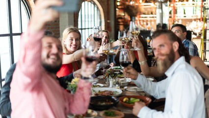Group of people enjoying a meal, raising glasses, and taking a selfie. Diverse group celebrating, smiling, and dining together in a lively restaurant setting. Friends at festive dinner taking selfie.