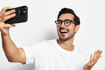 Happy smiling young man take selfie with smartphone, wearing stylish glasses and casual white...