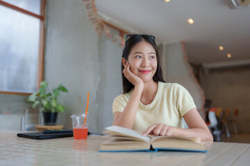 Young asian woman reading a book and relaxing with a refreshing drink in a modern cafe, enjoying...