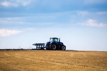 Fototapeta premium A modern blue tractor with an attached reversible plow with a husking roller is plowing a field on which the spring grain crop has just been harvested. Midsummer in central Ukraine.
