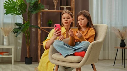 Young girl child spending evening time at home, girl sits in chair in the living room with mother and shopping online on smartphone, mom typing card number in app.
