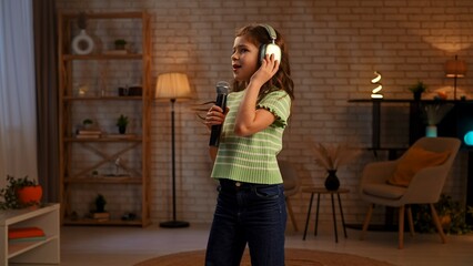Young girl child spending time at home, girl in living room listening music in headphones and singing in karaoke microphone, positive expression.