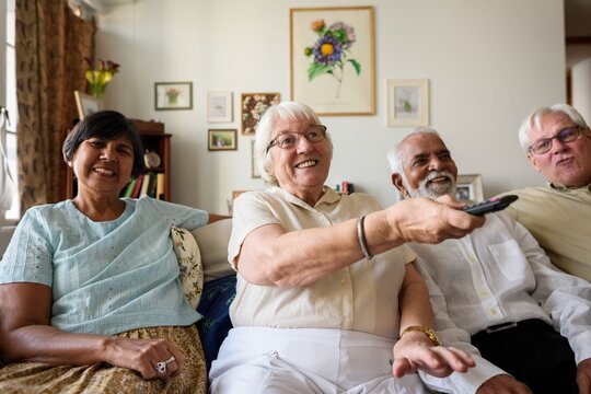 Group of seniors sitting on a couch watching TV. Diverse elderly friends enjoying time together on a couch watching television. Diverse elderly men and women have good time holding remote on a couch - Powered by Adobe