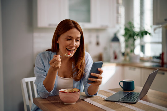 Happy woman using cell phone while eating healthy breakfast in kitchen.
