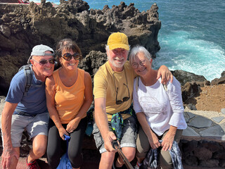 Four cheerful seniors along the sea coast looking at phone for selfie - Group of smiling pensioners enjoying nature and taking a walk together on a sunny day