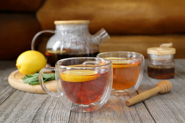 On an old wooden table against a log wall, in front of a tray with a glass teapot, lemon, mint and a glass jar of honey, there are two glass cups of hot black tea.