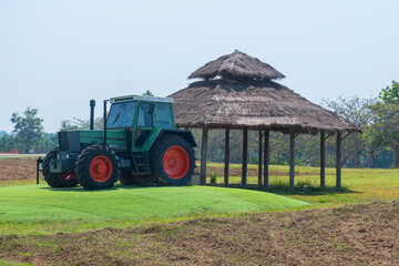 Green tractor parked beside rustic wooden hut with thatched roof on farmland in agricultural field, Chiang Mai, Thailand. Agriculture and farming concept