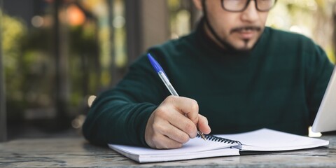 Man writing in notebook, focused on notes. Writing with pen, wearing glasses. Notebook and pen on table, writing and note-taking in progress. A man taking notes in notebook outdoor.
