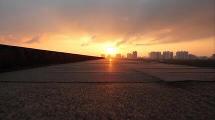 Photograph of a long, empty road with a city skyline in the background. the sky is a beautiful orange and yellow color, with the sun setting in the distance.