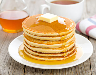 Delicious stack of fluffy pancakes topped with pat of butter and drizzled with syrup, served on white plate. warm cup of tea is in background, creating cozy breakfast scene