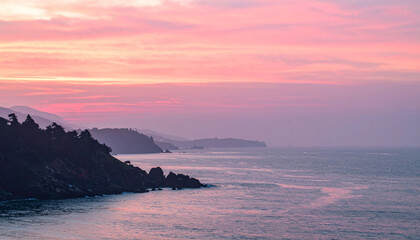 Fototapeta premium breathtaking coastal view featuring pink sunset over hazy waters, with silhouettes of distant hills and rocky shorelines creating serene atmosphere. soft colors evoke sense of tranquility