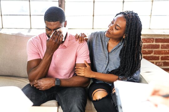 Comforting scene of two African American people on a couch. Man is carrying and the woman consoles him, showing empathy and support. Emotional support and empathy are key themes.
