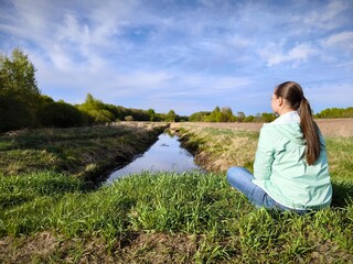 young woman is sitting alone in a meadow in early spring, against the background of a calm narrow...