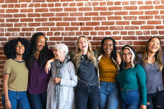 Diverse group of women smiling, standing against a brick wall. Women of various ethnicities, casual attire, joyful expressions, togetherness, unity. Diverse group of women united, standing together.