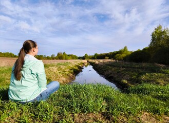 young woman is sitting alone in a meadow in early spring, against the background of a calm narrow...