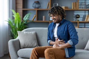 A young man sits on a sofa, experiencing stomach pain. He is holding his abdomen and appears to be...