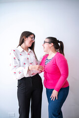 Professional businesswomen wearing glasses standing together, sharing lighthearted laughter near white wall during workplace break