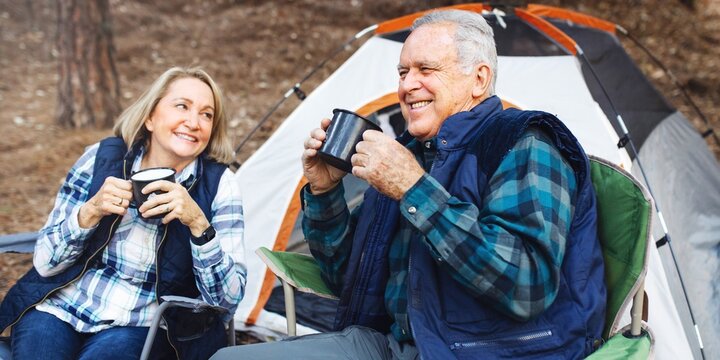 Elderly Caucasian couple camping, enjoy coffee at a tent. Happy elderly couple sit outside tent, drink coffee, outdoors camping. Elderly Caucasian couple drink coffee, happy outdoor nature camping