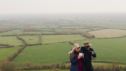 Elderly couple enjoying a scenic view. The couple, wearing jackets, stands on a hill. The couple admires the landscape, holding cups, enjoying the outdoors. Senior couple on a hike in the countryside.