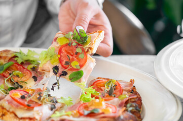 A person picks up a slice of pizza topped with fresh tomatoes, herbs, and greens from a plate,...