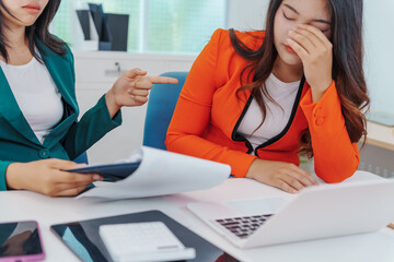 Two Asian businesswomen sit together at a desk, working professionally to solve problems. One...