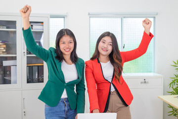 Two Asian businesswomen sit together at a desk, working professionally to solve problems. One...