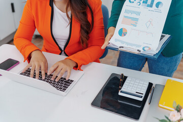 Two Asian businesswomen sit together at a desk, working professionally to solve problems. One...