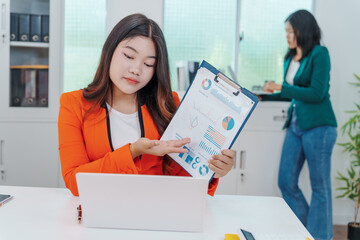 Two Asian businesswomen sit together at a desk, working professionally to solve problems. One...