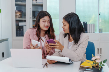 Two Asian businesswomen working together at a desk, focused and professional. They collaborate on...