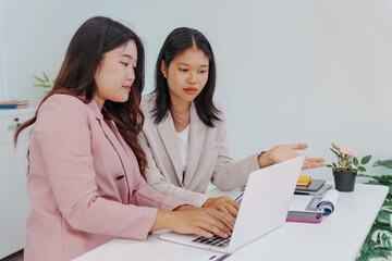 Two Asian businesswomen working together at a desk, focused and professional. They collaborate on...