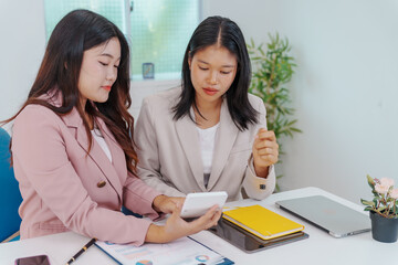 Two Asian businesswomen working together at a desk, focused and professional. They collaborate on...