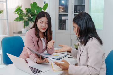 Two Asian businesswomen working together at a desk, focused and professional. They collaborate on...